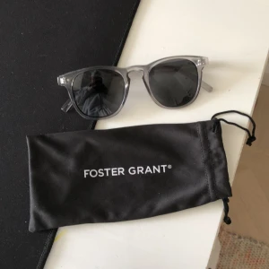 Grå transparenta solglasögon Foster Grant - Snygga solglasögon från Foster Grant med transparent grå båge och mörka glas. Klassisk rundad form som passar till de flesta stilar. Kommer med svart fodral med logga. Perfekta för soliga dagar och en clean look.