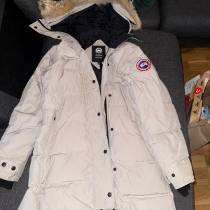 Beige dunjacka från Canada Goose M - Säljer en beige dunjacka från Canada Goose med päls på huvan och klassisk logga på ärmen. Jackan har lång passform, svarta knappar och dragkedja framtill. Insidan är svart och jackan är quiltad för extra värme. Perfekt för kalla vinterdagar.