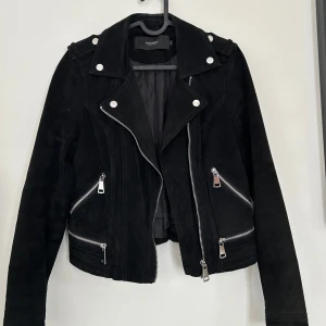 Svart mockajacka från Vero Moda - Snygg svart mockajacka från Vero Moda med coola silvriga dragkedjor och knappar. Jackan har bikerstil, lång ärm och flera fickor med dragkedja. Jackan är använd ett fåtal gånger och är i fint skick!