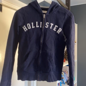 Hollister hoodie - Mörkblå hoodie från Hollister i storlek XS med vit broderad logga över bröstet. Tröjan har huva, långa ärmar och praktiska fickor framtill. Tillverkad i mjuk bomullsmix och stängs med dragkedja. Perfekt för chill dagar eller när du vill ha en sportig look. På bild 4 ser man att de har broderats in Californien som jag tagit bort, men råkat göra ett litet hål som man kan knappt se. Säljer för att den är för liten