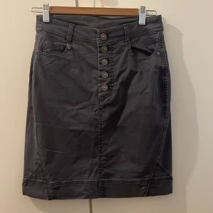 Grå kortkjol från House of Lola - Snygg mörkgrå kortkjol från House of Lola i jeansmaterial. Kjolen har fem dekorativa knappar framtill, klassiska fickor och bälteshällor. Passformen är normal och stilen är enkel men cool, perfekt för dig som gillar en stilren look. Syl XS men passar S. 