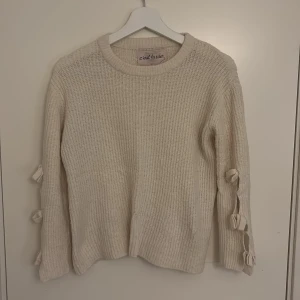 Kräm vit stickad tröja med rosetter - Superfin kräm vit stickad tröja från C'est la vie. Tröjan har långärmade ärmar och snygga rosettdetaljer längs båda ärmarna som ger en söt och trendig touch. Perfekt för dig som gillar mysiga och stilrena plagg med det lilla extra.