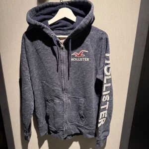 Blå hoodie med dragkedja från Hollister - Mörkblå hoodie från Hollister med broderad logga på bröstet och stort tryck på ena ärmen. Tröjan har huva med snörning, två fickor fram och dragkedja. Mjuk insida och klassisk streetstil – perfekt för chill dagar.