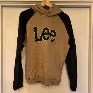 Beige och svart Lee hoodie med logga - Snygg hoodie från Lee i beige med svarta ärmar och stor Lee-logga framtill. Tröjan har huva med snörning och är tillverkad i mjuk bomullsmix. Perfekt för dig som gillar streetwear och vill ha en chill look.