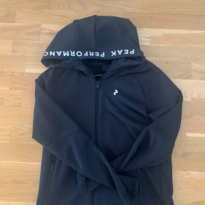 Svart hoodie Peak Performance - Svart hoodie från Peak Performance med vit logga på bröstet och text på huvan. Tröjan har dragkedja framtill, två fickor med dragkedja och lång ärm. Perfekt för chill eller träning, riktigt clean och sportig look. Inte andvänd alls mycket!