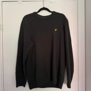 Mörkgrön stickad tröja från Lyle & Scott - Mörkgrön stickad tröja från Lyle & Scott med klassisk rund hals och gul broderad logga på bröstet. Tröjan är långärmad och har ribbade muddar vid ärmslut och nederkant. Perfekt för dig som gillar stilren och enkel design.
