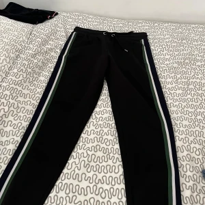 Svarta lacoste mjukisbyxor med ränder - Svarta lacoste mjukisbyxor med vita och gröna ränder längs sidorna. Byxorna har snörning i midjan och en sportig vibe, perfekta för chill eller träning. Sköna och stilrena med normal passform.