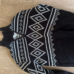 Svartvit stickad tröja med mönster - Svart stickad tröja med vita geometriska mönster och halv dragkedja vid halsen. Tröjan har hög krage och långa ärmar, perfekt för kalla dagar. Klassisk nordisk stil som sticker ut och är supermysig.