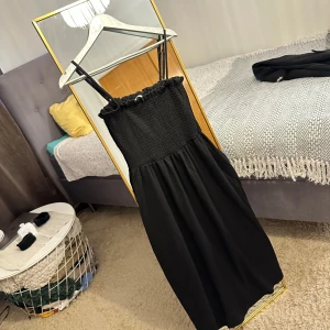 Svart långklänning från H&M - Svart långklänning från H&M med smockad överdel, smala axelband och volangdetalj upptill. Klänningen har en luftig kjol och är perfekt för dig som gillar enkel men stilren design. Materialet känns lätt och bekvämt.