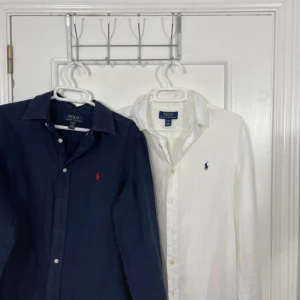 2 st linneskjortor Polo Ralph Lauren - Två klassiska linneskjortor från Polo Ralph Lauren, en i marinblått med röd broderad logga och en i vitt med blå logga. Båda har lång ärm, knappar framtill och avslappnad passform. Perfekta för en clean och stilren look. Köpta för cirkus 2000 tillsammans. Storlek 14-16 vilket är typ xs 