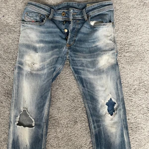Blå slitna jeans från Diesel - Säljer ett par blå jeans från Diesel med coola slitningar och hål på båda benen. Jeansen har klassisk femficksdesign, knappar i gylfen och snyggt tvättad denim med ljusa partier. Perfekta för en avslappnad streetstil.