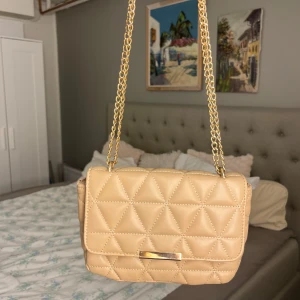 Beige axelväska från Glitter - Snygg beige axelväska från Glitter med quiltad design i triangelmönster. Väskan är i bra skick använd väldigt fåtal gånger. Tillverkad i syntetmaterial. 