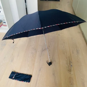 Mörkblå rutig paraply med fodral - Snyggt mörkblått paraply med rutiga detaljer i rött, blått, svart och vitt längs kanten och på remmen. Kommer med matchande fodral och har ett smidigt handtag med handledsrem. Perfekt att ha i väskan för regniga dagar.