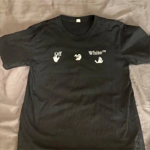 Svart Off-White t-shirt med regnbågstryck - Snygg svart t-shirt från Off-White med vit logga och handmotiv framtill. Baksidan har ett stort regnbågsfärgat tryck med pilar och text. Klassisk passform och rund hals. Perfekt för dig som vill sticka ut med streetwear-vibe. Nyskick storlek medium