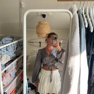 Grå omlottopp från NA-KD Reborn - Grå omlottopp från NA-KD Reborn i satinliknande material. Toppen har långa puffärmar, croppad passform och knytband i midjan. Oanvänd, bara provad.