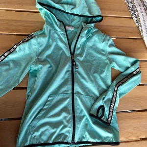 Turkos Träningskofta - Ljus turkos träningskofta från H&M Sport med huva och svarta detaljer. Jackan har hel dragkedja, fickor fram och texten 'YOU GO GIRL' längs ärmarna. Perfekt för aktiva dagar och träning. Materialet är lätt och känns skönt mot huden. Koftan är i stl 122/128 men passar modellen på bilden som vanligtvis har stl 146/152!
