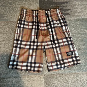 Burberry shorts - Snygga shorts från Burberry med klassiskt rutigt mönster i beige, svart, vitt och röda detaljer. De har elastisk midja med svart snörning och Burberry-logga på benet. Perfekta för sommaren och ger en clean och trendig vibe.