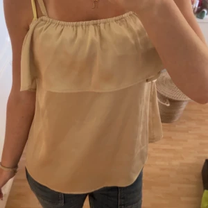 Champagnefärgad linnetopp i satin - Supersöt linnetopp i champagnefärgad satin med tunna axelband och volangdetalj upptill. Sval och glansig känsla, perfekt för varma dagar. Sitter löst och har en enkel, stilren look som passar till jeans eller kjol.