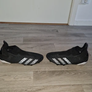 Adidas Predator fotbollsskor svart/vit - Säljer ett par Adidas Predator fotbollsskor i svart med vita detaljer och klassiska tre ränder på sidorna. Skorna har plösfri design, mönstrad ovandel och fasta dobbar för gräsplan. Tillverkade i syntetmaterial för bra passform och kontroll.