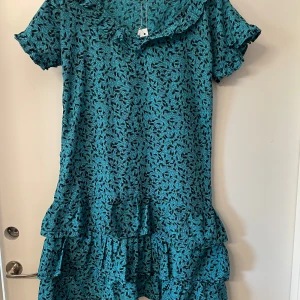 Turkos blommig kortklänning med volang - Supersöt turkos kortklänning med svart blommönster och volangdetaljer nertill och vid ärmslut. Klänningen har korta ärmar och en lätt oversized passform. Perfekt för dig som gillar färg och mönster!