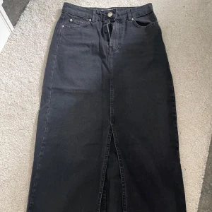 Svart lång jeanskjol med slits från BERSHKA - Säljer en svart lång jeanskjol med klassisk femficksdesign och snygg slits framtill från BERSHKA. Kjolen har knapp och dragkedja i midjan och är perfekt för dig som gillar en stilren och edgy look.