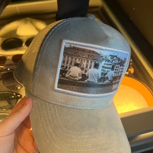 Grå keps från Sayless med foto-patch - Snygg grå keps från Sayless med mesh på sidorna och en stor patch framtill med svartvitt foto av tre personer vid en stadsmiljö. Kepsen har böjd skärm och justerbar baksida. Perfekt för dig som gillar streetstyle och unika detaljer. Helt ny!