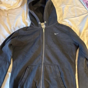 Svart Nike hoodie med dragkedja - Svart hoodie från Nike i storlek M med klassisk vit logga på bröstet. Tröjan har huva med snörhål, hel dragkedja framtill och två stora fickor. Tillverkad i mjuk bomullsblandning, perfekt för chill eller träning.