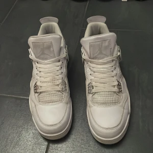 Nike Air Jordan 4 pure money - Säljer ett par Nike Air Jordan 4 Retro i helvitt skinn med silverdetaljer och klassisk mesh på sidorna. Skorna har snörning, rund tå och ikonisk Jumpman-logga bak. Perfekta för dig som vill sticka ut med stilrena sneakers.