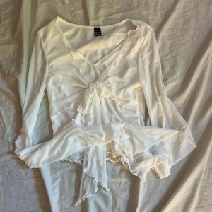 Vit omlottopp med volang från Shein - Superfin vit omlottopp från Shein med långa ärmar, volangdetaljer och knyt framtill. Tunn och flowy känsla, perfekt för lager-på-lager. Djup v-ringning och romantiska vibes med de vågiga kanterna.