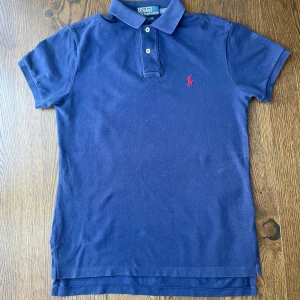 Marinblå pikétröja från Ralph Lauren - Klassisk marinblå pikétröja från Ralph Lauren med röd broderad logga på bröstet. Tröjan har korta ärmar, krage och två knappar framtill. Tillverkad i mjuk bomull och har en snygg, normal passform. Perfekt för en clean och stilren look. Pris kan diskuteras 