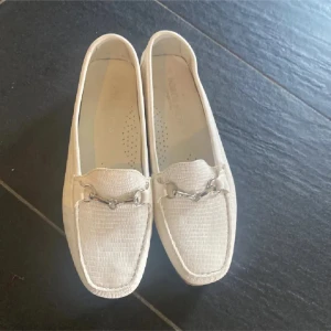 Beige loafers med metallspänne - Snygga beige loafers med diskret struktur i materialet och ett stilrent metallspänne framtill. Skorna har platt sula med rutmönster undertill och en klassisk, tidlös design. Perfekta för dig som gillar en clean och fräsch look. Tryck ej köp nu
