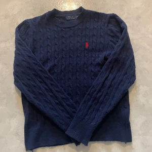 Mörkblå stickad tröja Polo Ralph Lauren - Kabelstickad mörkblå tröja från Polo Ralph Lauren med klassisk rund hals och röd broderad logga på bröstet. Inte använd så många gånger och väldigt bra skick
