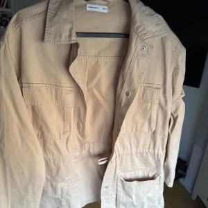 Beige overshirt från Perfect Jeans - Snygg beige overshirt från Perfect Jeans med flera praktiska fickor framtill och knappar. Jackan har dragsko i midjan för en schysst passform och är tillverkad i mjuk bomull. Perfekt att slänga över en hoodie eller t-shirt för en chill look.