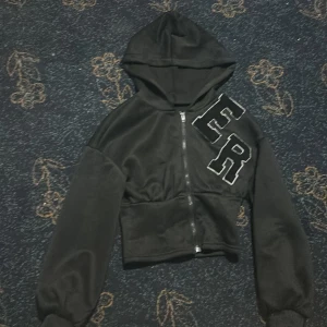 Svart croppad hoodie med dragkedja - Svart croppad hoodie med stora broderade bokstäver 'ER' på bröstet. Tröjan har huva, lång ärm och dragkedja framtill. Insidan är mjuk och fleece-liknande, perfekt för kyligare dagar. Oversized ärmar och bred mudd nedtill ger en trendig look.