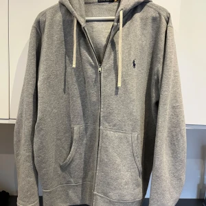 Grå hoodie från Polo Ralph Lauren - Snygg grå hoodie från Polo Ralph Lauren med dragkedja, huva med vita snören och klassisk blå logga på bröstet. Tröjan har känguruficka framtill och är tillverkad i mjuk bomull. Perfekt för en chill och stilren look.