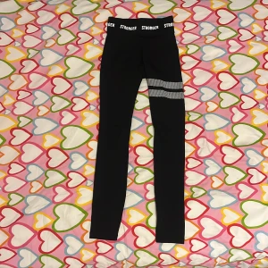 Svarta tights från Stronger XXS - Svarta tights från Stronger i storlek XXS med bred resår i midjan där det står 'STRONGER'. Byxorna har två vita prickiga ränder på ena benet och är tillverkade i stretchigt material med 75% återvunnen polyester och 25% elastan. Nypris är kring 700-800kr men jag säljer för endast 200kr!