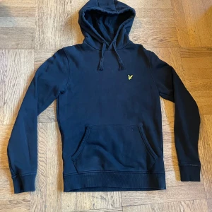 Svart hoodie från Lyle & Scott - Svart hoodie från Lyle & Scott med klassisk gul logga på bröstet. Tröjan har huva med snörning och en stor magficka framtill. Tillverkad i mjuk bomull, perfekt för chill dagar. Långärmad och med ribbade muddar för extra komfort.