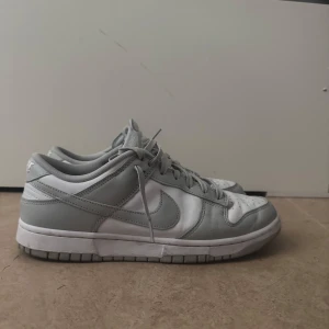 Nike Dunk Low i vitt och grått - Nike Dunk Low sneakers i en clean vit och ljusgrå färgkombination. Klassisk låg siluett med snörning, perforerad tåbox och tydlig Swoosh på sidorna. Ovandel i skinn och syntet, perfekt för dig som gillar stilrena och tidlösa sneakers.