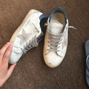 Vita sneakers från Golden Goose - Snygga vita sneakers från Golden Goose med grå stjärndetalj på sidan och grå snörning. Skorna har en mörkblå hälkappa och klassisk platt sula. Tillverkade i skinn med en cool, avslappnad vibe och retroinspirerad look. Allt og följer med.