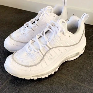 Nike Air Max 98 helvita sneakers - Snygga Nike Air Max 98 sneakers i helvitt med coola detaljer och synlig Air-sula. Skorna har en mix av mesh och syntetmaterial, rund tå och snörning. Perfekta för dig som vill ha en clean och sportig look med maximal komfort.