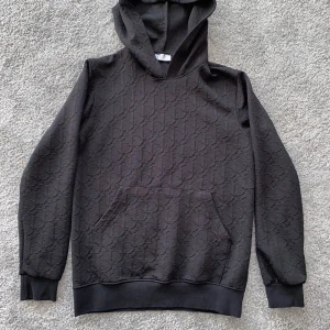 Dior oblique hoodie  - Säljer en svart hoodie från Dior i 100% bomull med snyggt, diskret Dior-monogram i hela tyget. Klassisk känguruficka framtill, huva och ribbade muddar. Perfekt för dig som gillar exklusiva streetwear vibes och vill sticka ut med stil.