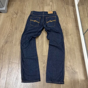 Nudie Jeans (Straight Fit) - Snygga mörkblå jeans från Nudie Jeans med klassisk rak passform och kontrastsömmar. Jeansen har fem fickor, orange broderi på bakfickorna och läderpatch med logga bak i midjan. Tillverkade i robust denim av bomull.