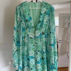 Blommig långklänning från Adoore - Superfin långklänning från Adoore i grönt och blått med blommigt mönster. Klänningen har v-ringning, långa puffärmar och knappar framtill. Perfekt för sommarens alla tillfällen och ger en romantisk vibe.