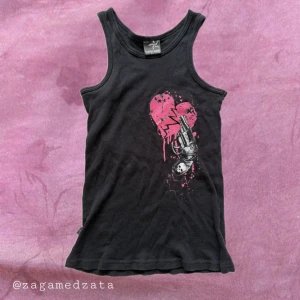 Svart ribbad linnetopp Darkside - Svart ribbad linnetopp från Darkside med tryck av ett rosa trasigt hjärta och en revolver på framsidan. Toppens material är stretchigt och sitter tight. Perfekt för dig som gillar alternativ stil och vill sticka ut med edgy detaljer.