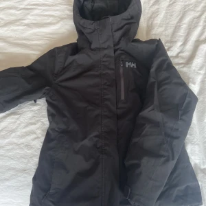 Svart skidjacka Helly Hansen - Svart skidjacka från Helly Hansen i storlek XS. Jackan har huva, dragkedja framtill och en bröstficka med dragkedja perfekt att förvara ett liftkort i. Jacka är supervarm men har ändå vädringsfunktioner så den passar båda kalla som lite varmare dagar. Den har ett praktiskt snölås och fleece i ryggen för ytterligare värme. 