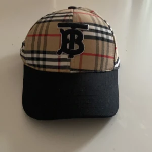 Burberry rutig keps med svart skärm - Snygg keps från Burberry med klassiskt rutigt mönster i beige, svart, vitt och rött. Framsidan har en svart broderad TB-logga och skärmen är helsvart. Kepsen är tillverkad i bomull och har en stilren och tidlös look.