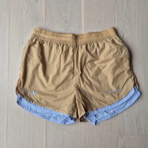 Adidas x Wales Bonner shorts beige/blå - Snygga sportiga shorts från Adidas x Wales Bonner i beige med ljusblå detaljer och broderade loggor. De har elastisk midja, kort passform och är perfekta för sommaren. Materialet är lätt och känns luftigt, perfekt för träning eller chill dagar.