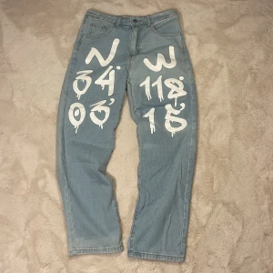 Ljusblå jeans med vit graffiti print - Unika ljusblå jeans med vit graffiti-inspirerad print framtill. Klassisk femficksmodell med raka ben och normal passform. Materialmix av bomull, viskos och polyester. Perfekta för dig som vill sticka ut med en streetstyle-look. Inte något använda.