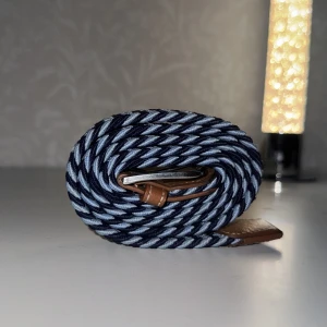 Randigt blått bälte med läderdetaljer - Snyggt randigt bälte i marinblått och ljusblått med bruna läderdetaljer vid spännet och änden. Bältet har ett flätat mönster och metallspänne, perfekt för att ge outfiten en cool touch.