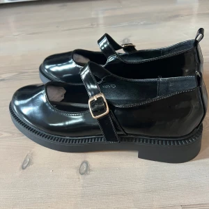 Svarta Mary Jane ballerinaskor C.M Paris - Helt oanvända. Glansiga svarta Mary Jane skor i lack från C.M Paris med rund tå, justerbar rem över foten och guldfärgat spänne. Skorna har chunky platt sula och är tillverkade i syntetmaterial med lackad finish. Perfekta för en trendig och stilren look.